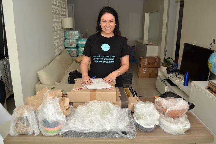 Foto que ilustra matéria com dicas de como fazer mudança organizada mostra a Personal Organizer Aline Delavy com papel e caneta na mão em meio a diferentes caixas com itens embalados nelas.