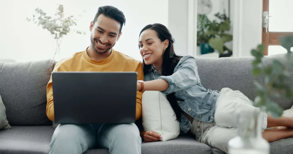 Imagem de um homem e uma mulher sorridentes sentados juntos em um sofá cinza, olhando para um laptop no colo do homem. A mulher aponta para a tela, ambos felizes e relaxados em uma sala de estar iluminada e aconchegante.