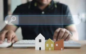 Imagem de uma pessoa usando um teclado de computador com três pequenas casas modelo em primeiro plano e uma sobreposição gráfica de barra de pesquisa transparente por cima