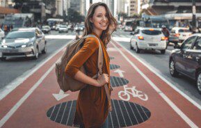 Imagem de uma mulher sorridente com uma mochila está no meio de uma ciclovia na Avenida Paulista, em São Paulo, cercada por carros e prédios altos em uma rua movimentada.