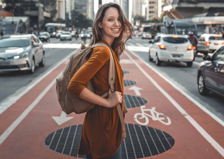 Uma mulher sorridente com uma mochila está no meio de uma ciclovia na Avenida Paulista, cercada por carros e prédios altos em uma rua movimentada.