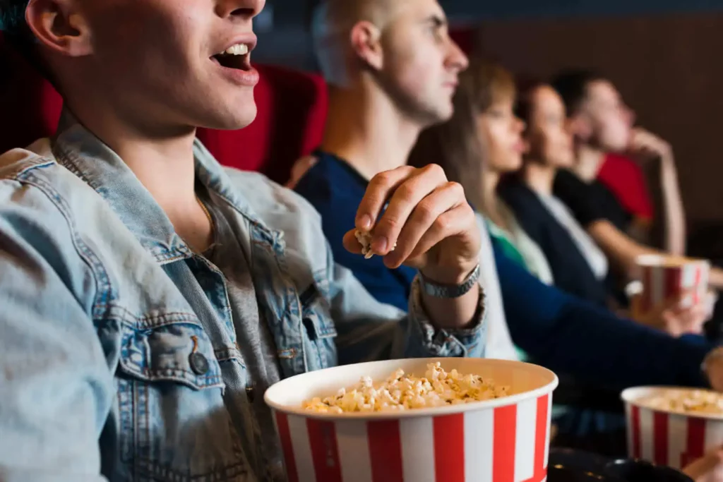 Imagem de um grupo de pessoas sentadas em uma sala de cinema, assistindo a um filme e comendo pipoca de baldes listrados, com foco em uma pessoa na frente usando uma jaqueta jeans.