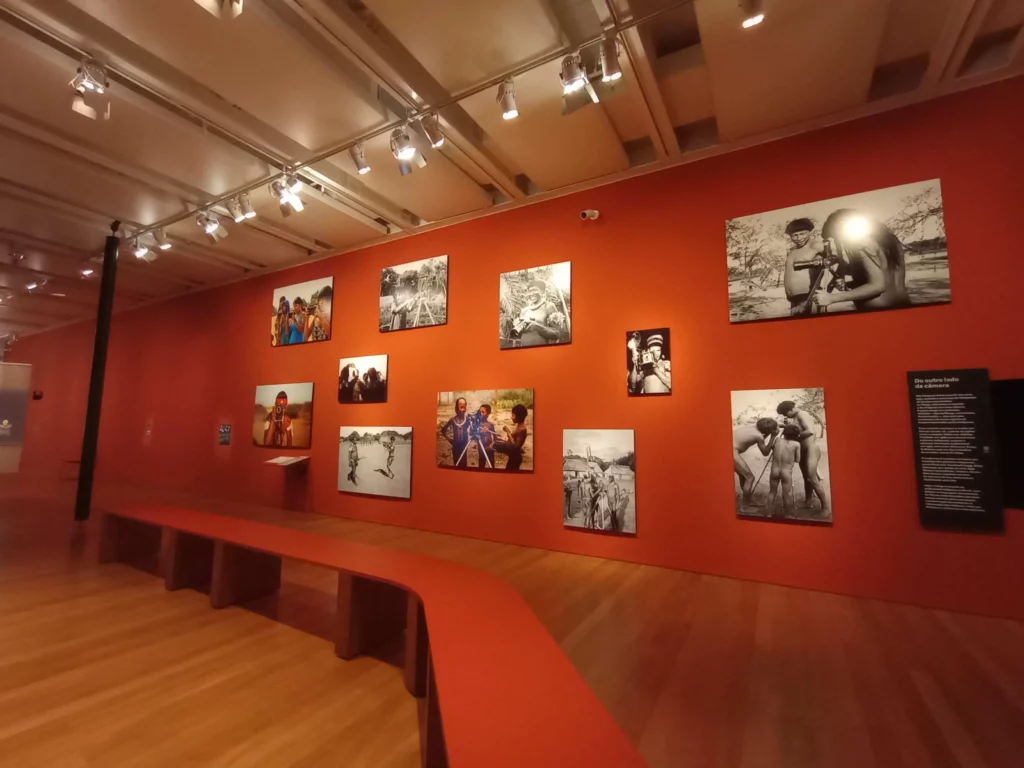 Imagem de uma galeria do Instituto Moreira Salles, na Avenida Paulista, com paredes vermelhas adornadas com fotografias coloridas e em preto e branco de pessoas em várias poses. Um longo banco vermelho alinha-se no piso de madeira, enquanto holofotes iluminam as obras de arte.