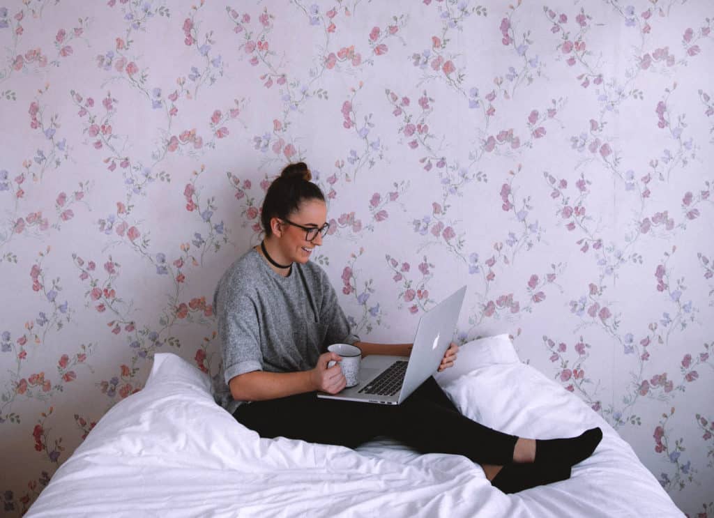 Mulher em almofada branca usando um laptop, com papel de parede ao fundo.