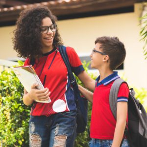Duas crianças sorridentes, com mochilas e cadernos, vestindo camisetas vermelhas, caminham perto de uma área verde. A mais velha tem cabelos cacheados e óculos; a mais nova olha para elas enquanto conversam.