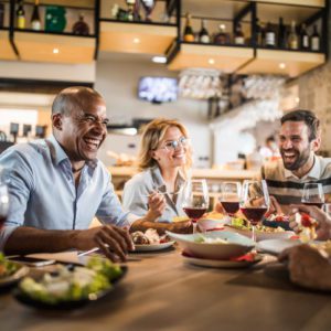 Cinco adultos estão sentados à mesa de um restaurante, rindo e apreciando uma refeição juntos. Pratos de comida e taças de vinho estão à sua frente, e o ambiente é animado e amigável.