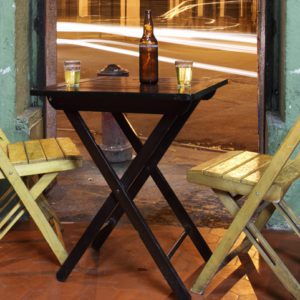 Duas cadeiras de madeira estão de frente para uma pequena mesa escura com uma garrafa, dois copos e um cinzeiro, colocados em uma porta aberta para uma rua com luzes desfocadas de carros à noite.