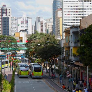 Uma movimentada rua urbana com modernos arranha-céus, dois ônibus em faixas exclusivas, pedestres caminhando pelas calçadas e lojas ao longo da rua, tudo isso em um cenário com céu limpo.