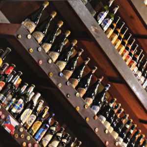 Fileiras de garrafas de cerveja variadas expostas em prateleiras de madeira inclinadas, com vários rótulos coloridos e tampas de garrafa visíveis, criando um ambiente de bar ou loja vibrante e organizado.