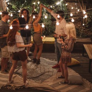 Um grupo de jovens adultos dança e socializa ao ar livre à noite, cercados por luzes de corda. Alguns seguram bebidas e conversam, enquanto outros dançam em tapetes estampados, criando uma atmosfera animada e aconchegante.
