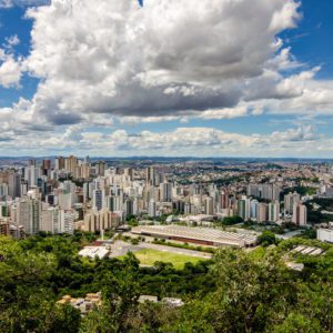 Uma vista panorâmica de uma grande cidade com muitos edifícios altos, cercada por vegetação e árvores em primeiro plano, sob um céu cheio de nuvens dramáticas.