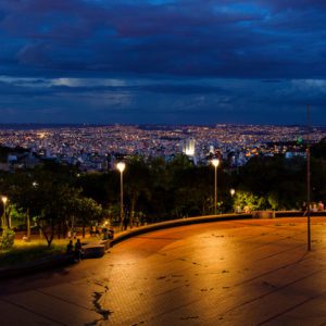 Uma paisagem urbana ao entardecer, vista de um parque com postes de luz brilhantes e pessoas espalhadas. A cidade abaixo é iluminada por muitas luzes sob um céu noturno dramático e nublado.