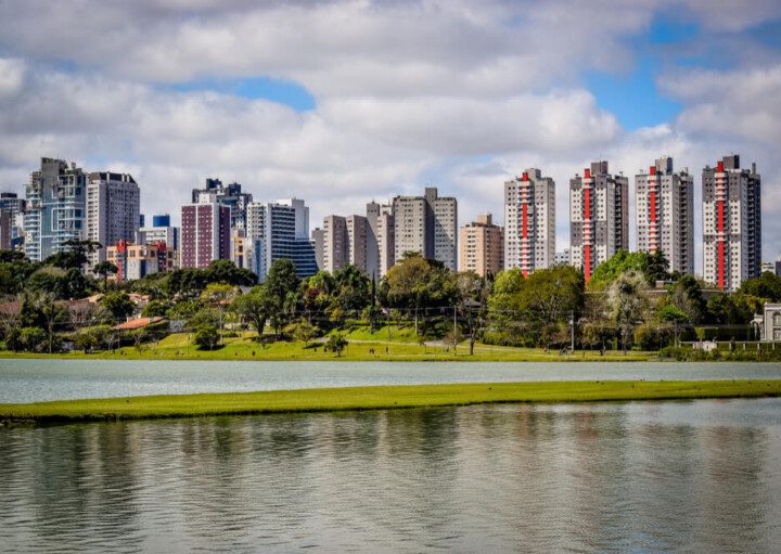 Foto que ilustra matéria sobre qualidade de vida em Curitiba mostra uma panorâmica do Parque Barigui, com um espelho d’água em primeiro plano, árvores no meio da imagem e altos prédios ao fundo