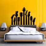 Um quarto moderno com uma parede amarela vibrante apresenta um adesivo preto com vários headstocks de guitarra acima da cabeceira da cama, adicionando um toque de decoração musical. Duas mesas de cabeceira com luminárias ficam de cada lado da cama.
