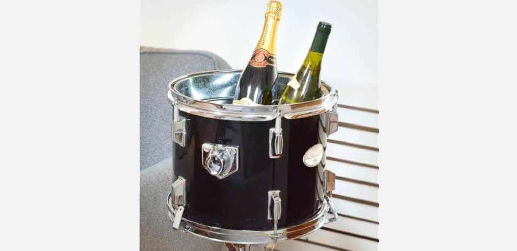 Um tambor preto transformado em balde de gelo contém uma garrafa de champanhe e uma garrafa de vinho branco, acrescentando um toque de decoração musical ao lado de um sofá cinza.