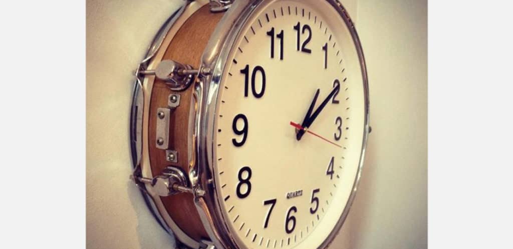 Um relógio de parede com mostrador branco e números pretos está situado dentro de um tambor, misturando elementos de um instrumento musical e de um relógio — perfeito para quem busca decoração musical em uma parede branca.
