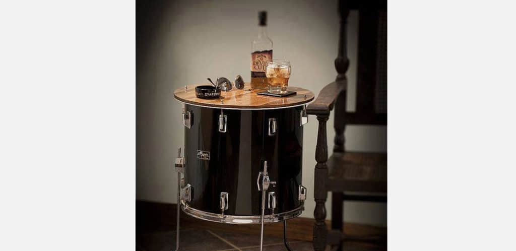 Um tambor transformado em mesa lateral contém uma garrafa de rum, um copo com gelo e um molho de chaves ao lado de uma cadeira de madeira escura, adicionando um estilo de decoração musical único ao espaço.