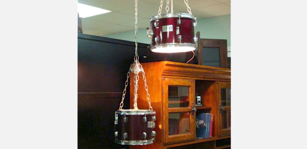 Duas luminárias pendentes feitas de cascos de tambor reaproveitados com correntes trazem um toque de decoração musical, suspensas acima de um armário de madeira com portas de vidro e livros dentro.