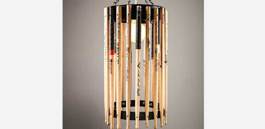 Uma luminária pendente feita de baquetas de madeira verticais dispostas em formato cilíndrico ao redor de uma lâmpada central, com anéis de metal preto na parte superior e inferior — perfeita para adicionar um toque de decoração musical ao seu espaço.