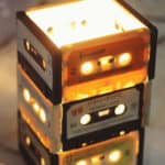 Um abajur feito de fitas cassete empilhadas e iluminadas brilha com luz quente, trazendo um toque de decoração, musicalidade e criatividade nostálgica a qualquer espaço.