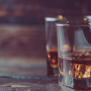 Dois copos de u&iacute;sque com cubos de gelo sobre uma superf&iacute;cie de madeira, com um copo em primeiro plano e o outro desfocado ao fundo. A cena tem um ambiente acolhedor e r&uacute;stico.