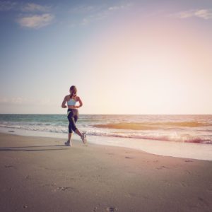 Uma pessoa corre ao longo de uma praia arenosa perto da costa ao p&ocirc;r do sol, com ondas calmas e um c&eacute;u claro ao fundo.