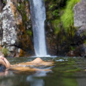 Uma pessoa flutua de costas em uma piscina cristalina na base de uma cachoeira, cercada por penhascos rochosos e plantas verdejantes. O cen&aacute;rio &eacute; tranquilo e natural.
