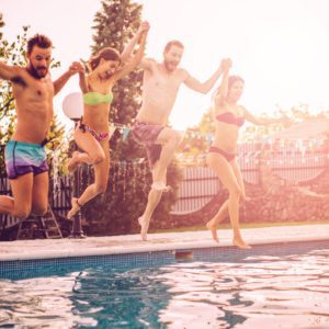 Quatro amigos em trajes de banho est&atilde;o de m&atilde;os dadas e pulando alegremente em uma piscina juntos em um dia ensolarado, com &aacute;rvores e decora&ccedil;&otilde;es vis&iacute;veis ao fundo.