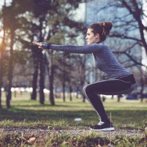 Uma mulher com roupa esportiva realiza um exerc&iacute;cio de agachamento ao ar livre em um parque ao nascer ou p&ocirc;r do sol, com &aacute;rvores e um pr&eacute;dio ao fundo. Uma garrafa de &aacute;gua est&aacute; no ch&atilde;o ao lado.