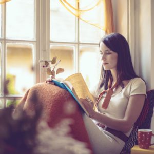 Uma mulher est&aacute; sentada perto de uma janela, enrolada em um cobertor, lendo um livro. A luz suave do sol se filtra pelas cortinas transparentes, e uma caneca vermelha repousa ao lado dela, criando uma atmosfera aconchegante e tranquila.