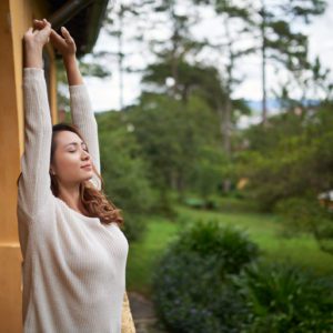 Uma mulher de cabelos longos estica os bra&ccedil;os acima da cabe&ccedil;a e fecha os olhos, em p&eacute; ao ar livre, ao lado de um pr&eacute;dio amarelo com um jardim tranquilo e &aacute;rvores ao fundo.