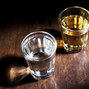 Dois copos de shot sobre uma superf&iacute;cie de madeira; um cont&eacute;m um l&iacute;quido transparente, provavelmente &aacute;gua ou vodca, e o outro cont&eacute;m um l&iacute;quido marrom-dourado, possivelmente u&iacute;sque. Ambos os copos projetam sombras na luz.