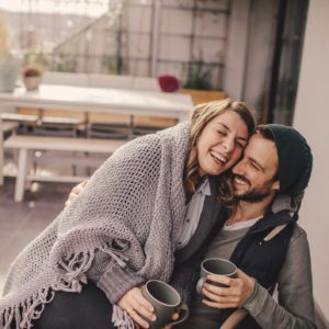 Um casal feliz sentado bem juntinho em um p&aacute;tio, enrolado em um cobertor cinza e segurando canecas, sorrindo e apreciando a companhia um do outro. O fundo mostra uma mesa, cadeiras e plantas ao ar livre.