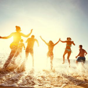 Seis pessoas correm e pulam no oceano ao p&ocirc;r do sol, espirrando &aacute;gua enquanto aproveitam a praia juntas. O sol brilha forte atr&aacute;s delas, criando um cen&aacute;rio acolhedor, alegre e cheio de energia.