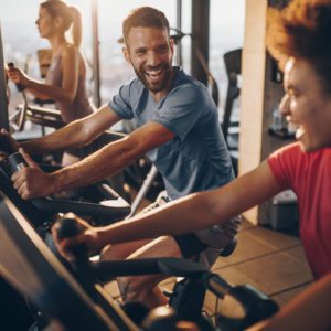 Duas pessoas, um homem e uma mulher, sorriem e conversam enquanto se exercitam em bicicletas ergom&eacute;tricas em uma academia bem iluminada. Outra pessoa pedala ao fundo, perto de uma grande janela.