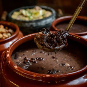 Uma concha retira uma por&ccedil;&atilde;o de feijoada de uma grande panela de barro. Ao fundo, veem-se outras panelas de barro com comida.