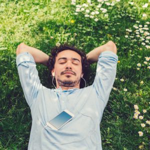 Um homem est&aacute; deitado na grama verde com pequenas flores brancas, com os olhos fechados e as m&atilde;os atr&aacute;s da cabe&ccedil;a, ouvindo m&uacute;sica com fones de ouvido conectados a um smartphone apoiado no peito.