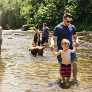 Um grupo de adultos e crian&ccedil;as caminha em um rio raso em um dia ensolarado, com um homem ajudando uma crian&ccedil;a a caminhar. Um cachorro est&aacute; na &aacute;gua e &aacute;rvores preenchem o fundo.