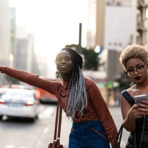 Duas jovens est&atilde;o em uma rua da cidade; uma est&aacute; sorrindo e apontando animadamente enquanto a outra, usando &oacute;culos, olha para o celular com fones de ouvido. Tr&acirc;nsito e pr&eacute;dios altos s&atilde;o vis&iacute;veis ao fundo.