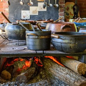 Panelas pretas de ferro fundido com cabos de madeira repousam sobre um fog&atilde;o a lenha r&uacute;stico, com toras e brasas acesas embaixo. Utens&iacute;lios de cozinha pendurados na parede, e uma cesta &eacute; vis&iacute;vel ao fundo.