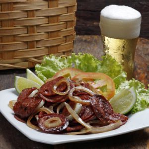 Um prato de lingui&ccedil;a grelhada com cebola salteada, alface, tomates fatiados e fatias de lim&atilde;o em um prato branco, ao lado de um copo de cerveja espumoso, sobre uma mesa de madeira r&uacute;stica com uma cesta de vime ao fundo.