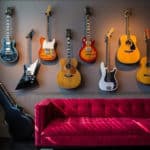 Uma coleção de guitarras elétricas e acústicas está pendurada em uma parede cinza acima de um sofá de veludo vermelho vibrante, adicionando um toque de decoração musical. A luz do sol cria padrões de sombras no sofá, enquanto um estojo de guitarra repousa no chão ao lado.