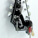 Um conjunto de chaves de casa está pendurado nas cravelhas de um headstock preto de guitarra elétrica, montado em uma parede branca e reutilizado como porta-chaves — um toque de decoração musical criativo.