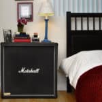 Um amplificador de guitarra Marshall serve como mesa de cabeceira única neste musical de decoração, coberto com uma luminária azul, livros, uma foto emoldurada e um vaso de flores ao lado de uma cama vestida com roupa de cama branca e um cobertor vermelho.