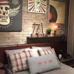 Um quarto aconchegante com uma parede de tijolos aparentes, decoração musical com pôsteres, uma estante com livros e decoração, almofadas xadrez e uma almofada cinza "Let It Be" na cama. Um telefone vintage no criado-mudo.