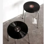 Dois bancos com discos de vinil como assentos adicionam um toque de decoração musical. Com pernas de metal e assentados sobre um piso de concreto perto de uma parede branca, estes bancos conferem um estilo único a qualquer espaço.