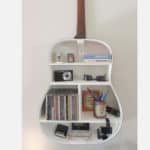 Uma prateleira de parede branca em formato de violão traz uma decoração musical ao seu espaço, exibindo livros, câmeras, óculos de sol, canetas em um pote, CDs, fitas cassete e outros itens pequenos em uma parede lisa de cor clara.