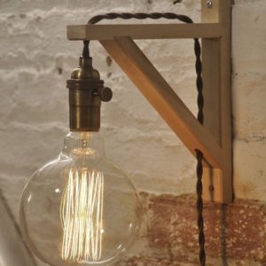 Uma l&acirc;mpada vintage exposta, com filamento vis&iacute;vel, pende de um suporte de madeira em uma parede de tijolos texturizados. Um fio marrom torcido corre ao longo do suporte, dando &agrave; lumin&aacute;ria um visual industrial.