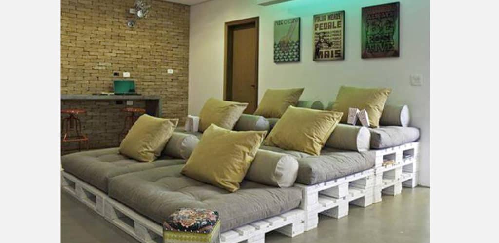 Um aconchegante cinema em casa com três poltronas em estilo sofá, feitas de pallets de madeira branca, cobertas com almofadas cinza e grandes travesseiros bege. As paredes apresentam alvenaria e obras de arte emolduradas.