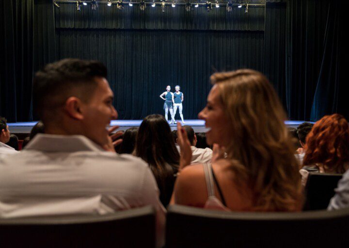 Casal observa fim de espetáculo em um teatro com casa cheia em São Paulo. Ao redor, todos aplaudem e o casal ri, dando a ideia de que gostou do show